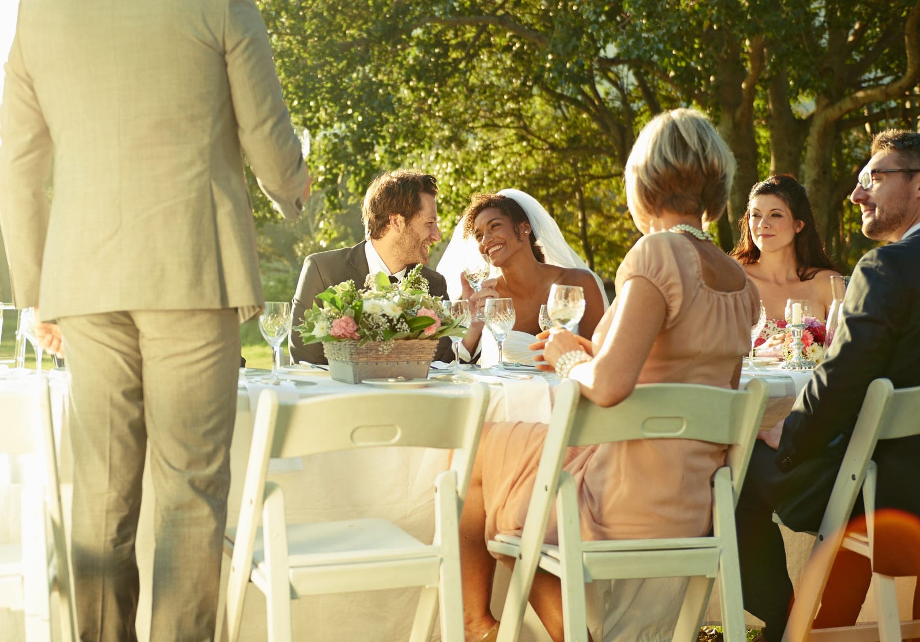 A newly married couple laughing together at their sunlit outdoor wedding reception, guests seated around the table